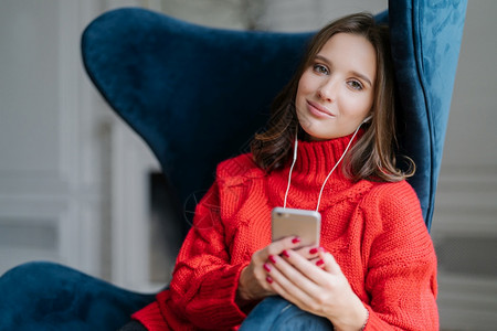 红色耳机成年少女在耳机中享有自由时间和流行音乐拥有现代智能电话穿红色编织毛衣坐在舒适的扶手椅上使用应程序检查新的更情况背景