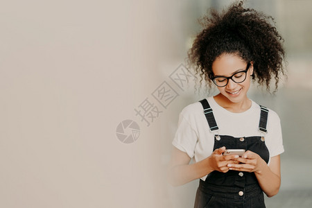 穿着时装的美籍女使用手机发送短信与wif连通检查电子邮件箱站在白墙附近空着间图片