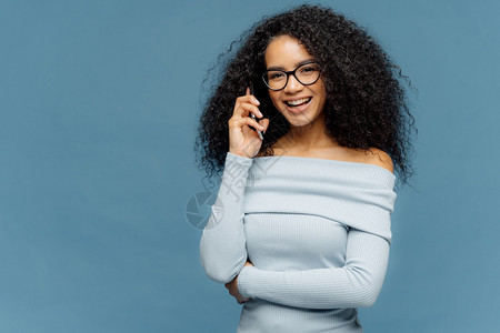 很高兴非洲女能享受电话交谈保持一只手在腰上使用现代技术穿戴眼镜和轻蓝跳跃器站在工作室空间你的口号图片