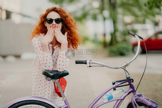 在户外拍摄华丽的红发女双掌盖着嘴部穿红色美甲着时装服戴和墨镜在自行车附近摆布免费骑自行车图片