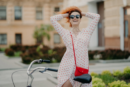 微笑着的女人在城市骑自行车保持双手在头上感觉自由戴太阳眼镜和暑期服装站在自行车附近独享乐趣引导健康的生活方式穿越街道图片