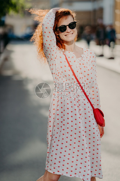 喜悦的年轻女士在街上漫步长发红穿时装墨镜周末享受阳光明媚的一天在模糊街道背景上装扮心情良好时间充裕图片