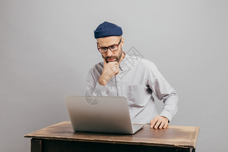 男自由职业者在互联网上读到新闻仔细看笔记本电脑观看录像坐在旧桌子上穿白衬衫和头盔隔着灰墙进行离图片