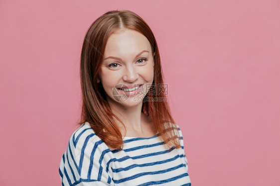 长得开心的棕色头发女人侧面镜头直发健康的皮肤愉快笑容穿着条纹毛衣与粉红色背景隔绝情感概念图片