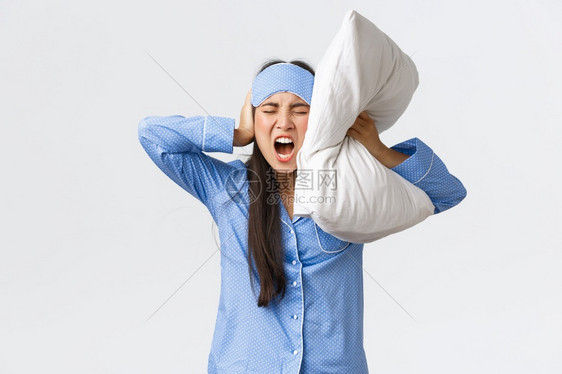 愤怒的亚洲女孩穿着睡衣和面具躺在床上用枕头闭耳朵大声叫喊因为吵得睡不着邻居在晚上通过聚会抱怨烦人的声音愤怒亚洲女孩穿着睡衣和面具图片
