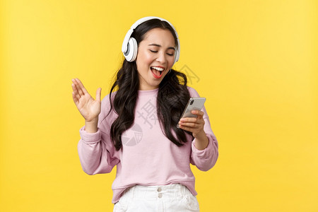 欢笑快乐和无忧虑的亚裔女孩在耳机上唱卡拉ok阅读手机的歌曲站立黄色背景人们的情感生活方式休闲和美丽概念欢笑快乐和无忧的亚裔女孩在图片