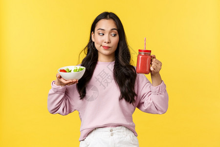 人们的情感健康生活方式和食物概念好奇可爱的亚洲女孩在看冰淇淋拿着沙拉和饮料选择早餐吃什么站着黄色背景人们的情感站在黄色背景图片
