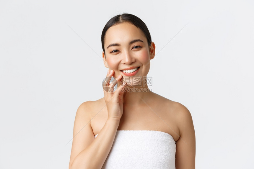 穿着毛巾的美丽年轻女站在浴室的毛巾微笑着快乐的相机触摸干净的皮肤在浴室施用皮肤护理产品照顾身体白色背景站在浴室的毛巾中美丽年轻女图片