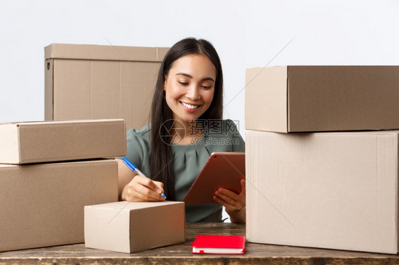 小企业主创和电子商务概念微笑着作为女商人的小企业主在有订单的箱子上书写货运地址的网上店主在有订单的箱子上书写货运地址在数字平板上图片