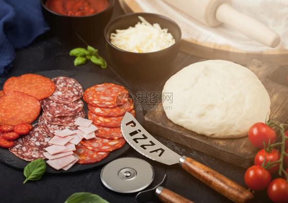 准备用生面粉烤番茄比萨饼沙拉米辣焦索切轮机新鲜西红柿和黑桌上的烤面包碗盘配奶酪和番茄糊图片