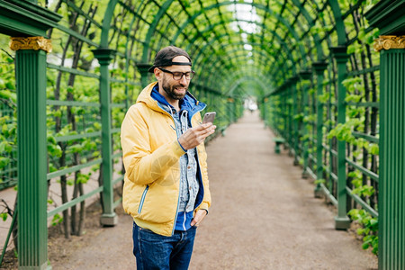 长胡子和的流行男身着厌食牛仔裤和帽站着专注地看他的智能手机将其隔绝在绿拱门之上图片