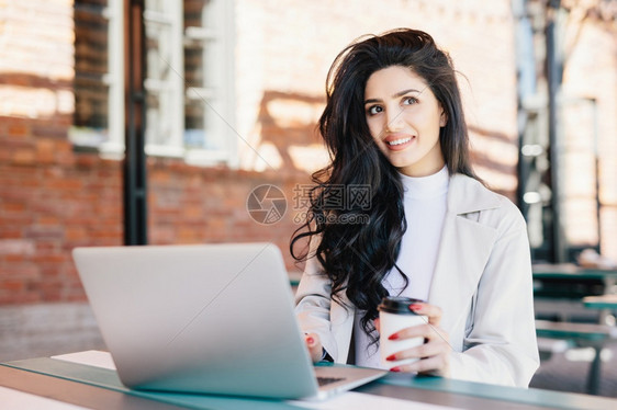 穿着长豪华头发的时髦黑女人穿着优雅的外衣在户咖啡厅做笔记本工作时带着梦想节日的深思熟虑表达方式向远处看去图片