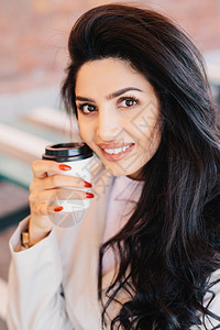 年轻褐色女眉健康皮肤和红指甲将咖啡放在镜头中笑得令人愉快地看图片