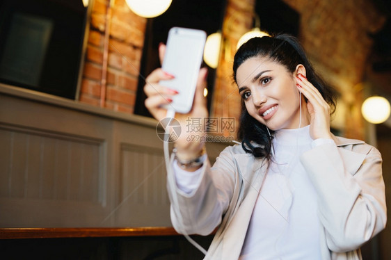 深色头发皮肤健康眼睛明亮手握智能机的有魅力妇女在咖啡馆休息时自拍听音乐或书籍图片