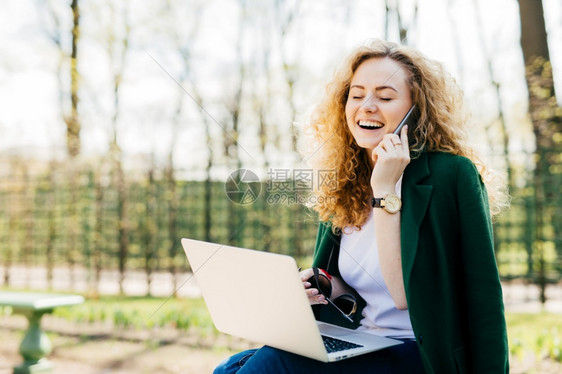 穿着绿色夹克的金发漂亮美女坐在公园户外讲着智能电话闭上眼睛很高兴听到她的男朋友读着笔记本电脑上的精灵书图片