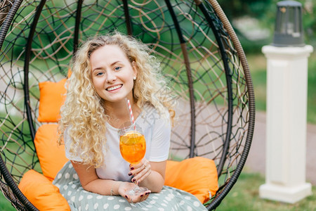有卷毛的可爱发年轻女拿着橙子鸡尾酒杯在露天椅子上摆姿势表示积极与对话者交谈愉快图片