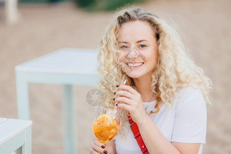美丽的年轻笑女长着卷发轻牙色笑容沙滩上的小礼服喝夏季新鲜鸡尾酒穿成白衬衫休息愉快人夏天生活方式概念图片