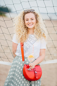 穿着T衬衫和裙子的小袋拿着冰的鸡尾酒站在海滩上的网球附近享受良好的休息图片