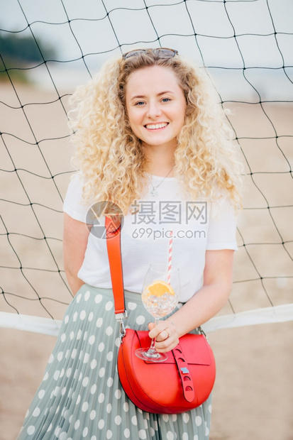 穿着T衬衫和裙子的小袋拿着冰的鸡尾酒站在海滩上的网球附近享受良好的休息图片
