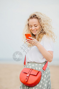 正面的年轻女使用现代智能手机使用愉快的表达方式在线购物携带明亮的小袋子在海边有户外散步人技术积极的概念图片