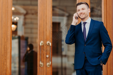 穿着优雅高贵西装的英俊年轻男老板肖像有移动电话对在咖啡馆门口站立在与合作伙伴见面后休息图片