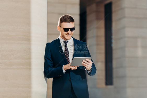 年轻时尚的男银行家研究未来的银行协议使用现代触摸垫阅读互联网的重要信息时戴太阳镜和奢华的正规礼服笑得高兴图片
