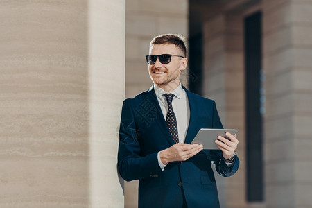 英俊年轻男雇主的肖像拥有数码平板电脑与无线互联网连接佩戴太阳镜和正式西装下班后有空闲时间背景图片
