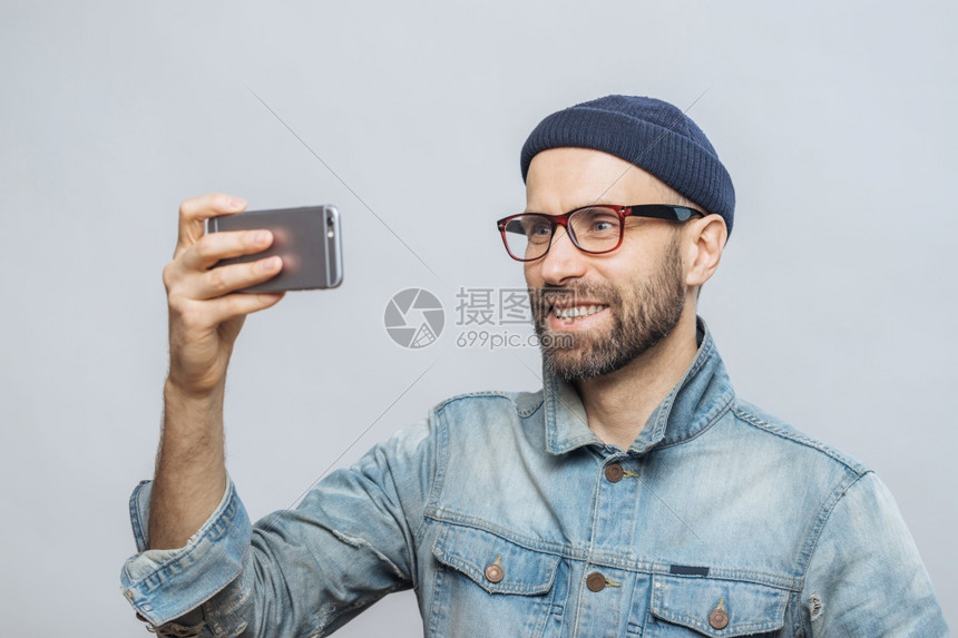 笑着的男在智能手机镜头上装快乐的表情拍摄自己的照片穿戴牛仔夹克和眼镜与白色背景隔绝图片
