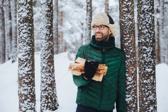 穿着绿色夹克的快乐胡须男子站在树旁拿着木柴看似置身事外开心的表情与积雪森林背景相对立图片