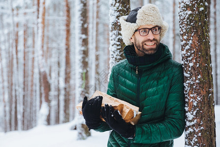 长胡子的年轻男戴着温暖的帽子和绿色夹克拿着一堆木柴看快乐的一旁注意到些东西比如冬天和寒冷的气梦想中的有吸引力人在森林里度过时光图片