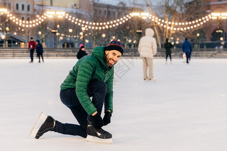 穿着绿色大衣的户外男子镜头滑冰鞋的带看着快乐的相机站在美妙的冰环上装饰着园林有积极的生活方式人休闲和冬季的概念图片