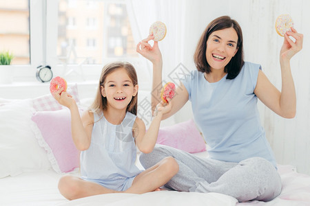 穿着睡衣的笑母亲和女儿早上心情很好拿着甜圈早餐吃点睡卧室化妆图片