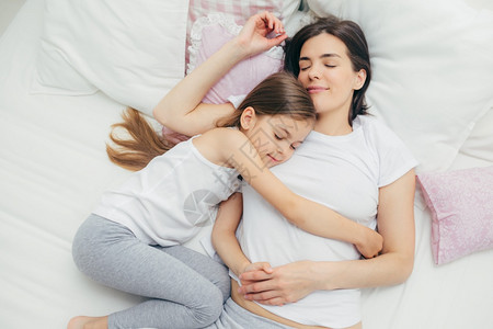 快乐的困睡母亲和可爱的女儿在床上拥抱做个愉快的梦躺在白睡衣上有良好的关系互相照顾图片