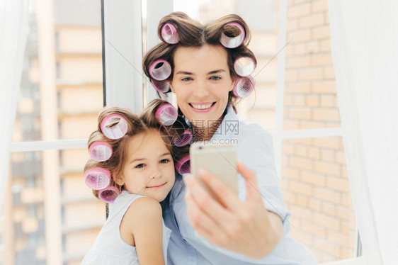 室内的一张照片以实在的表情看着妈和她的女儿做卷发风格在现代手机中装扮自拍准备嘉年华或派对图片