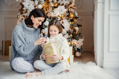 美丽的女孩和母亲肖像一起坐在温暖的地毯上拿着这个盒子享受盛装的新年树背景图片