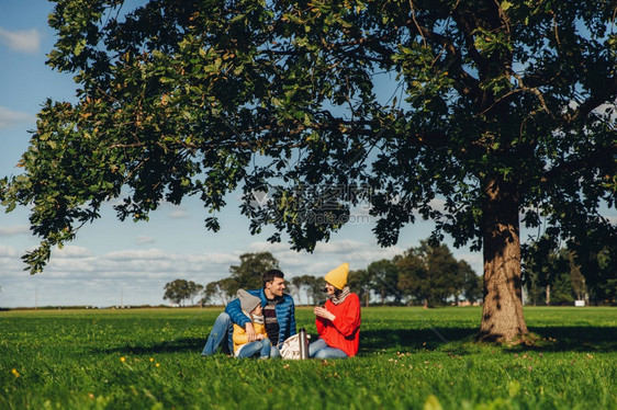 快乐的家庭有秋天野餐坐在绿草地上喝热茶相互交流享受平静的气氛仰慕蓝天和美丽的风景图片