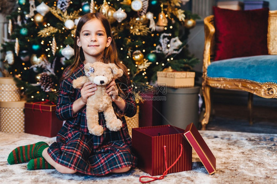 可爱的小女孩坐在地板上旁边是装饰新年树包的礼物很高兴从圣达克莱斯里拿起泰迪可爱的小孩子在圣诞节时会收到礼物奇迹和节日的概念图片