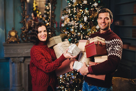 夫妇在新年或圣诞树附近站立持有许多礼物忙于节日准备有积极的表情男女青年准备庆祝图片