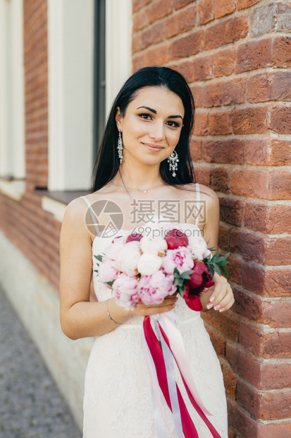 美丽的黑发女外貌迷人戴耳环穿白色婚纱着美丽的花束站在砖墙上图片