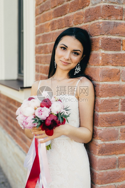 迷人的女新娘穿着白色婚纱拿花束站在砖墙附近有迷人的外貌图片