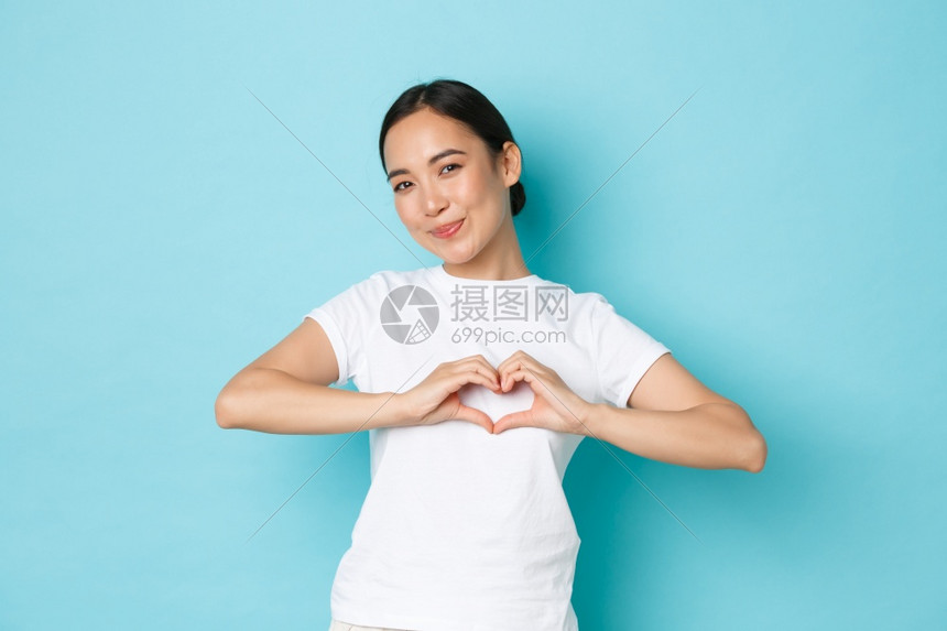 穿着白色t恤的可爱亚洲女孩穿着白色t恤对人表示同情达关心和温柔的感情展示心姿态在镜头面前笑站在蓝色背景上立穿着白色t恤的可爱亚洲图片
