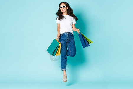 时装美和生活方式概念时装美丽和生活方式概念时装无照料的亚洲女在太阳眼镜中拿着购物袋和笑在商场散步轻蓝背景中跳起来图片