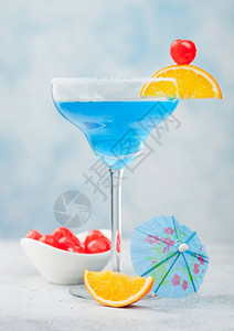 蓝色环礁夏季鸡尾酒在玛格丽塔杯中配有甜美鸡尾酒樱桃和橙色切片蓝桌底有雨伞图片