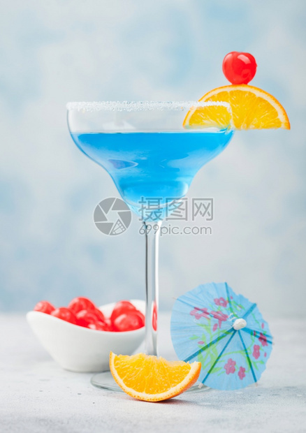 蓝色环礁夏季鸡尾酒在玛格丽塔杯中配有甜美鸡尾酒樱桃和橙色切片蓝桌底有雨伞图片