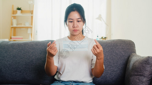 身戴保护面罩的沮丧女商人坐在沙发上家中客厅的沙发上当时社会不安定地呆在家中和自我隔离时间的流行病科罗纳毒概念图片