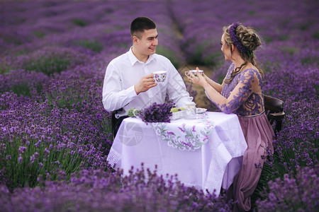 年轻夫妇坐在桌椅上熏衣草地喝咖啡或茶穿紫色裙子和花圈的妇女与男人一起在户外年轻夫妇坐在桌椅上熏衣草地喝咖啡或茶穿紫衣服和花圈的妇图片