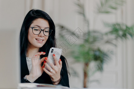 快乐的黑发女商人使用智能手机佩戴眼镜向伙伴发送短信从事项目工作在场所与舒适的内地抗争图片