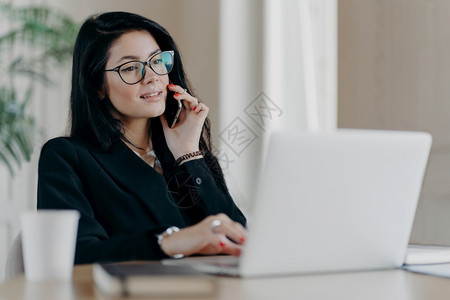 自信认真的女商人有电话交谈在笔记本电脑前摆姿势戴光学眼镜进行视力矫正在工作场所摆姿势使Resrach工作正式穿衣服图片