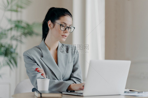 严重女创业者戴眼镜黑头发梳成马尾商业项目集中在笔记本电脑上穿着灰色正规服装使用网络资源信息图片