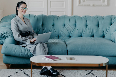 媒体营销公司官方网站的年轻繁荣女执行官在媒体营销公司正式网站上使用现代膝型计算机使用无线互联网在舒适的内阁工作坐在舒适的沙发上与图片